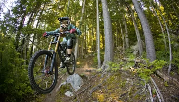 An adult woman rides off a rock on her downhill mountain bike on a singletrack trail near Nelson, B.C., Canada. She is riding a modern downhill mountain bike and wears a full face helmet, goggles and kneepads.